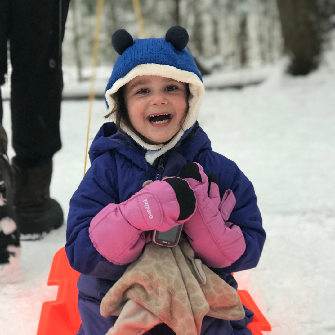 A little girl bundled in a snowsuit, hat and mittens smiles joyfully while being pulled on a sled